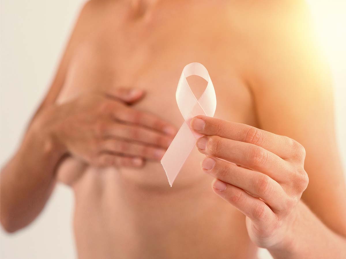 Mid section of a topless woman holding a pink ribbon and covering her breasts.